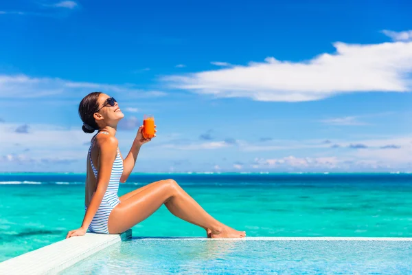 Foto msotra Mulher saudável de verão bebendo suco detox de cenoura para uma dieta equilibrada, perda de peso e um estilo de vida de biquíni. Garota feliz curtindo o sol e o bronzeado à beira da piscina de um hotel tropical. - bronzeamento, bronzar