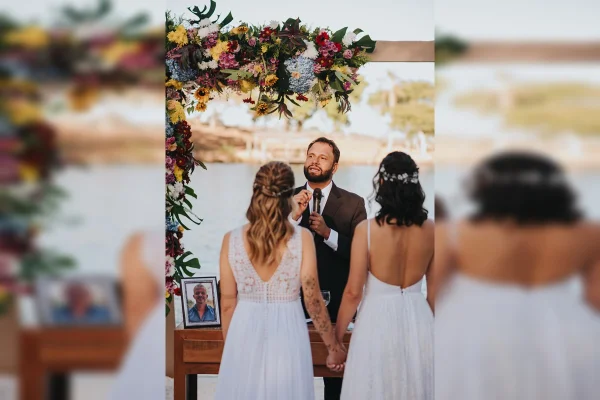 Imagem colorida de um casal de noivas, usando vestido branco, com o Pastor Henrique Vieira ao fundo. Ele usa um terno preto