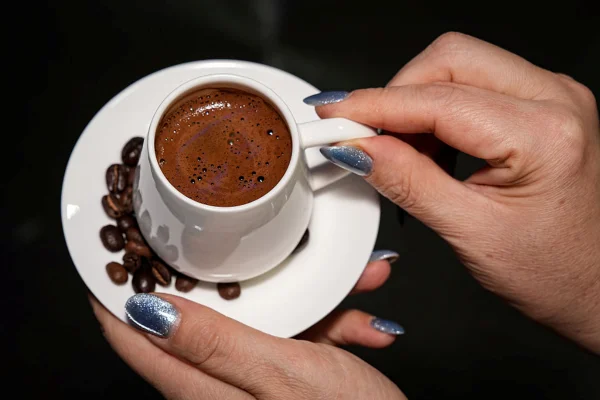 Foto mostra mão de mulher com esmalte prateado segurando uma xícara pequena de café e um píris com grãos de café