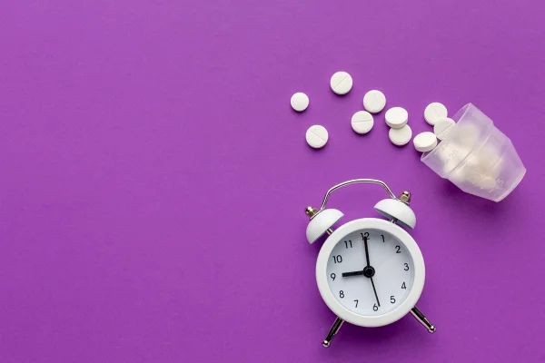 Foto colorida de fundo roxo, um despertador no canto inferior direito e comprimidos de cor branca - Metrópoles.