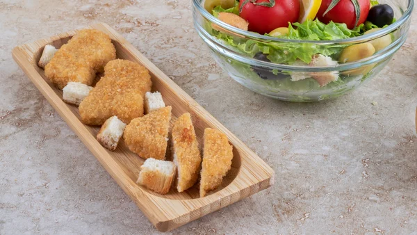 Foto colorida de nuggets de peixe disposto em tábua de madeira e salada ao lado