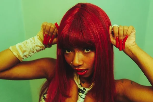 Foto colorida de uma mulher negra com cabelo vermelho. Ela faz uma pose mostrando as unhas vermelhas e longas, próximas do cabelo - Metrópoles