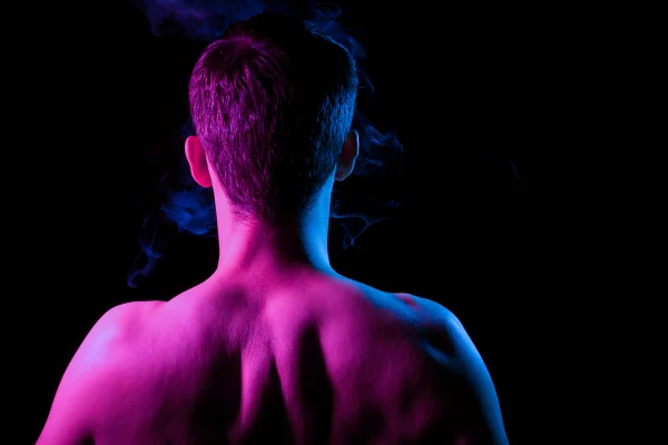 Close-up da nuca, costas e ombros com músculos tensos de uma cabeça de cabelo curto de um homem com barba por fazer, nu sem roupas, iluminado em azul e rosa em um fundo preto isolado com fumaça - Homem com condição rara vê o mundo cor de rosa ao ter orgasmos. Entenda