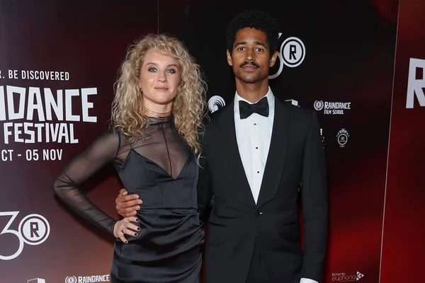Foto colorida de Alfred Enoch e Mona Godfrey em premiação, no tapete vermelho - Metrópoles