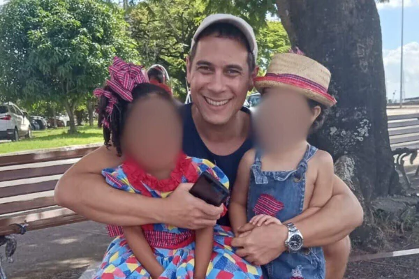Homem branco, de boné, sorrindo em parque, durante o dia. Ele abraça, com cada um dos braços, um menino e uma menina com roupas de festa junina - Metrópoes