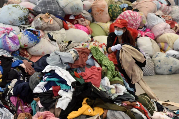 Mulher com máscara de proteção e boné mexendo em sacolas de roupas descartadas
