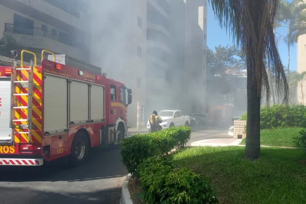 Quadra residencial, com prédio de paredes beges e vidros escuros, tomada por fumaça; há um caminhão de bombeiros na cor vermelha e branca parado; ao longe, há uma caminhonete branca estacionada; em primeiro plano, à direita, há um jardim, com plantas verdes e o tronco de uma árvore - Metrópoles