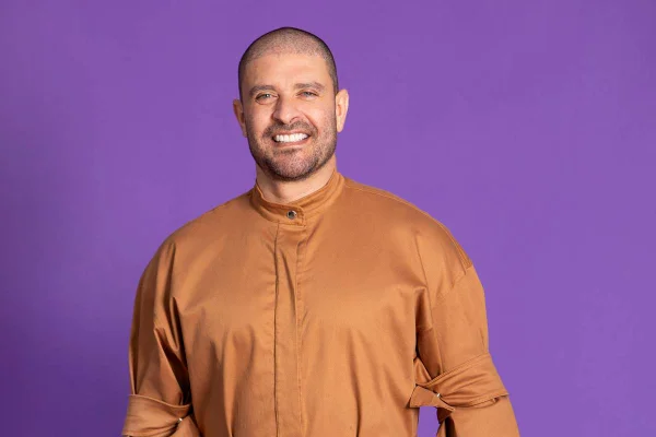 Cantor Diogo Nogueira vestido em macacão caramelo posa sorrindo em fundo roxo - metrópoles