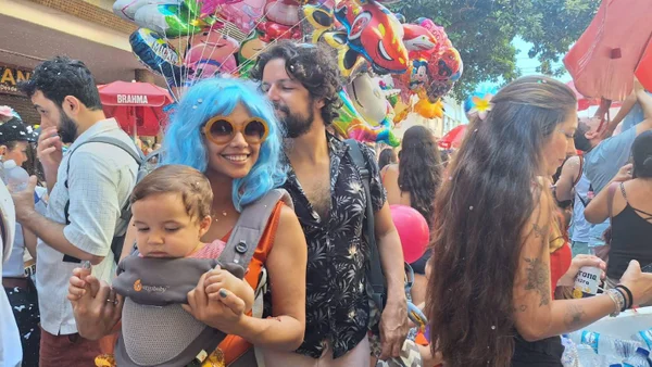 Foto colorida de mulher com peruca azul e óculos escuro carregando um bebê com diversas pessoas ao fundo durante bloco de Carnaval em São Paulo - Metrópoles