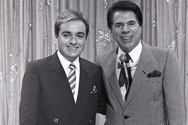 Foto antiga em preto e branco do apresentador Gugu e Silvio Santos, também apresentador e dono do SBT. Eles aparecem sorrindo e abraçados - Metrópoles