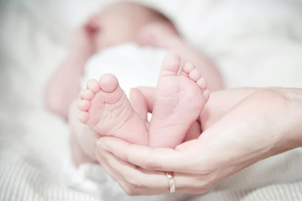 Imagem colorida de um bebê vestindo fraldas brancas e deitado sobre um lençol branco. Uma mão com uma aliança dourada segura os pés dele
