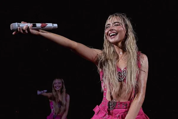 Foto colorida de Sabrina Carpenter em show, segurando microfone e com roupa rosa - Metrópoles
