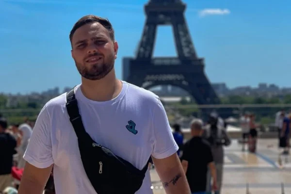 Imagem mostra Renan, que é um rapaz branco, de cabelos curtos e barba, com camiseta branca em frente à torre Eiffel - Metrópoles