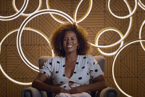 mulher preta com cabelo cacheado vestida em macacão branco de bolas pretas sorri sentada - metrópoles