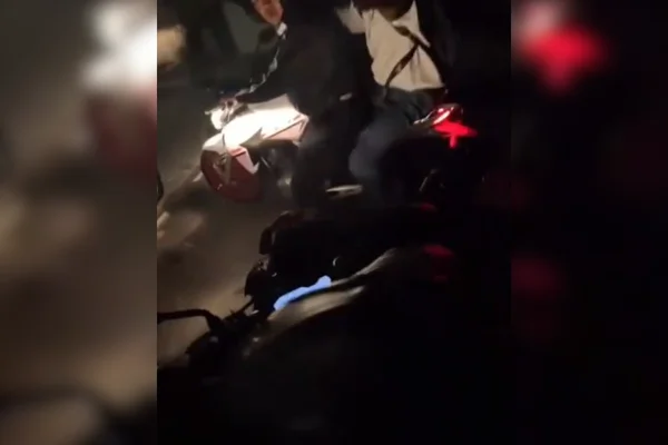 Imagem colorida de dois homens em uma motocicleta abordando outro homem durante a noite - Metrópoles