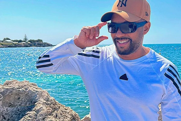 O cantor Salgadinho posa de boné, óculos escuros e blusa branca na praia - Metrópoles