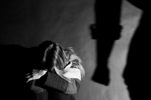 Imagem preto e branco de mulher acuada com cabeça apoiada nos joelhos, e sombra de mão masculina com punho fechado. Imagem conceito para violência contra mulher. - Metrópoles