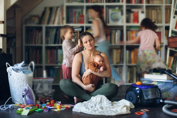 foto colorida de uma sala brinquetoteca com uma mãe cansada no centro e o filhos correndo