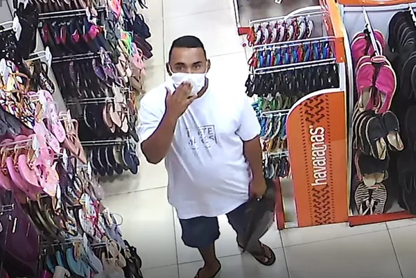 Homem com camisa branca e com a mão em máscara. Ele está dentro de loja de chinelos