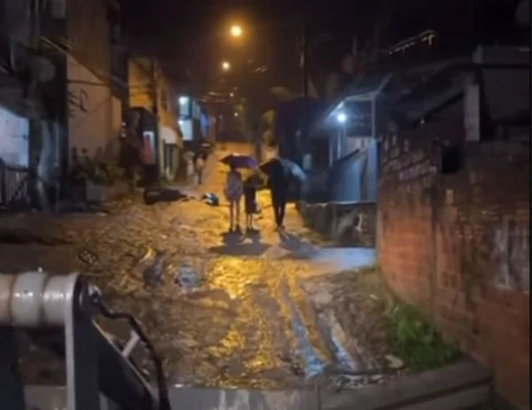 Foto colorida mostra Vila Sahy durante chuva forte. Duas pessoas aparecem caminhando com guarda chuva em viela cheia de lama