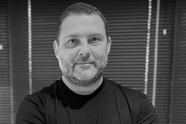 Imagem em preto e branco mostra homem branco com barba rala e rosto redondo. Ele está olhando para a câmera e veste uma camiseta preta - Metrópoles