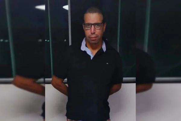 Homem de cabelo curtos e óculos veste camisa polo azul escura e cruza os braços atrás das costas