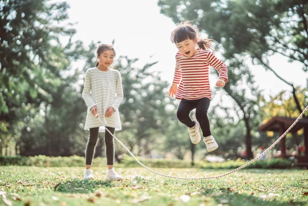 Foto colorida de duas meninas brincando de pular corda em área verde - Metrópoles