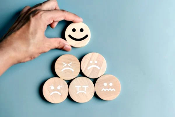 Mão de homem colocando uma carinha feliz de madeira próximo a outras carinhas de diferentes emoções de madeira formando uma pirâmide em fundo azul claro - Metrópoles