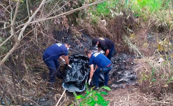 três homens retiram um corpo envolvido em um saco preto de dentro da mata