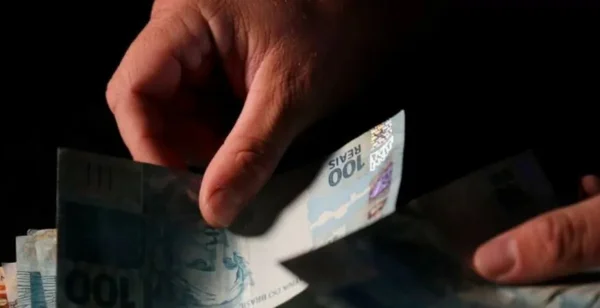 Mãos manuseando na penumbra notas de 100 reais - Metrópoles
