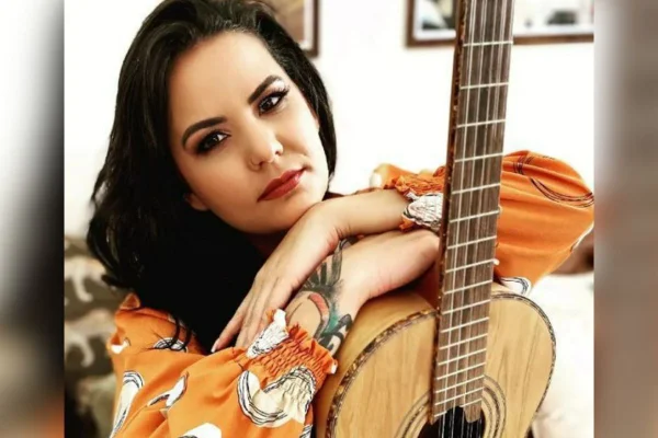 Imagem colorida mostra a cantora sertaneja Nanda Ferraz, uma mulher branca, de cabelos castanho escuros, apoiada em um violão - Metrópoles