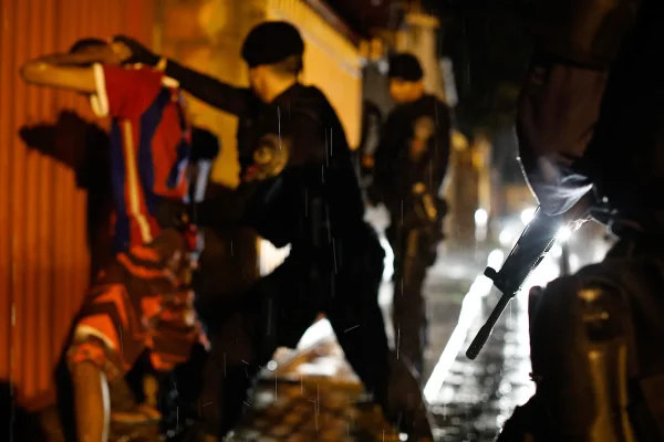 Imagem colorida, em luz baixa, de dois policiais abordando um homem e o colocando contra a parede