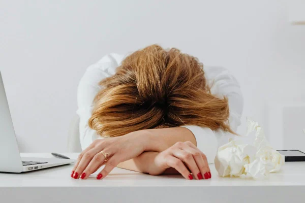 Mulher sentada em cadeira, debruçada sobre a mesa, com as mãos à frente da cabeça baixa, em claro sinal de cansaço, representando pesquisa sobre depressão e distúrbios do sono entre servidores da Educação e da Saúde estadual em SP