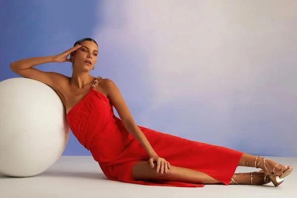 Mulher sentada no chão, apoiada em bola branca. Ela usa vestido vermelho assimétrico com fenda - Metrópoles