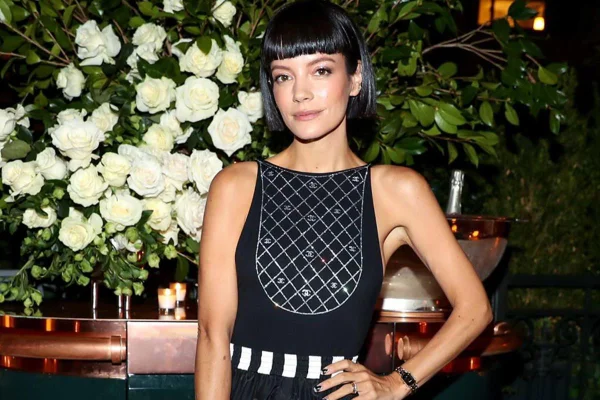 Foto colorida de Lily Allen. Ela é uma mulher branca, com cabelo chanel preto e usa vestido preto. Ao fundo, flores brancas - Metrópoles