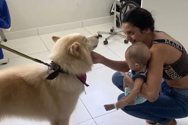 Foto colorida de mulher com bebê no colo fazendo carinho em um cachorro peludo