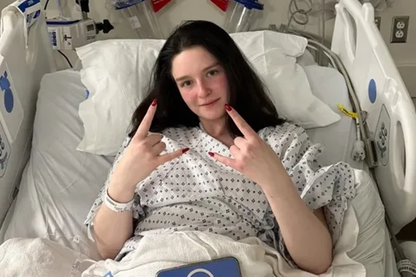 Foto colorida de mulher jovem de cabelos pretos, fazendo sinais com as mãos, enquanto está deitada a cama do hospital - Metrópoles.