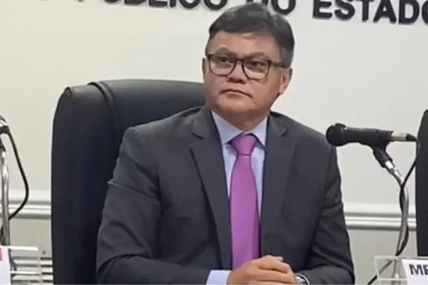 Homem com traços orientais, sem barba, óculos, trajando paletó da cor grafite, camisa social azul clara e gravata lilás clara, sentado em cadeira de couro prato, com mãos cruzadas, sobre uma mesa - Metrópoles