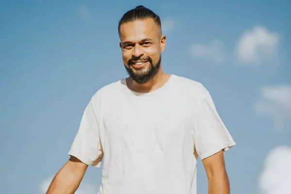 Foto colorida do ator Pierre Bittencourt. Ele está de blusa branca e sorrindo. Ele tem coque no cabelo preto e barba rala - Metrópoles