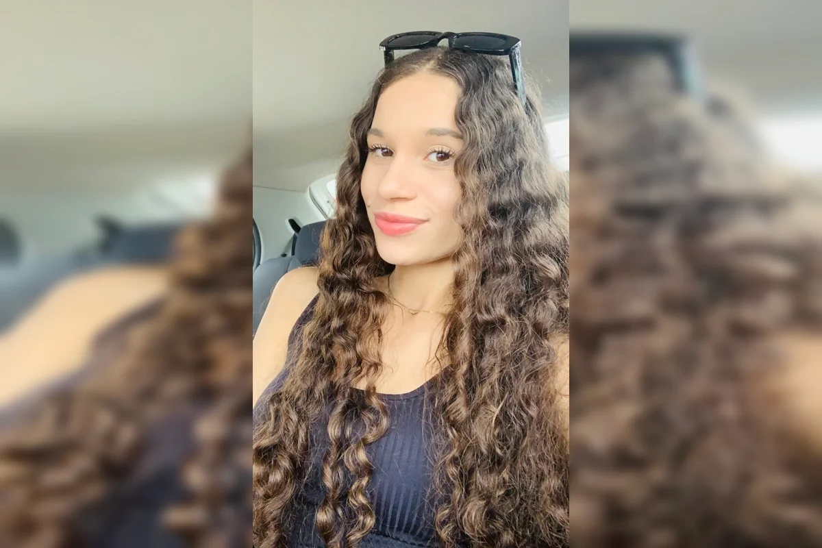 A jovem possui uma pinta bem característica na bochecha direita, próxima ao canto da boca, e cabelos castanhos escuros, longos e cacheados A jovem possui uma pinta bem característica na bochecha direita, próxima ao canto da boca, e cabelos castanhos escuros, longos e cacheados