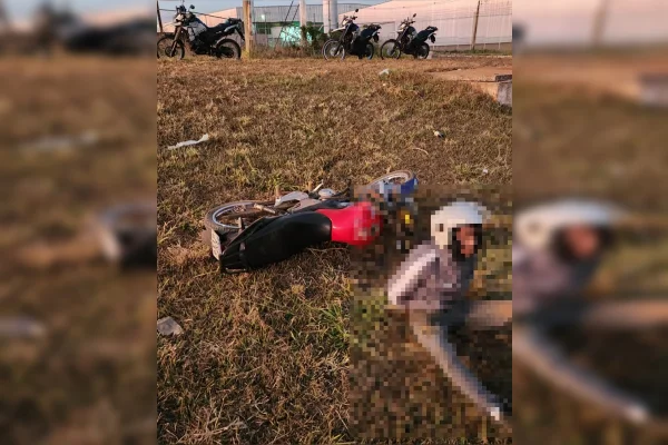 Imagem colorida de um homem sentado em uma grama, usando capacete. Atrás dele, há uma moto preta e vermelha, caída sobre a grama