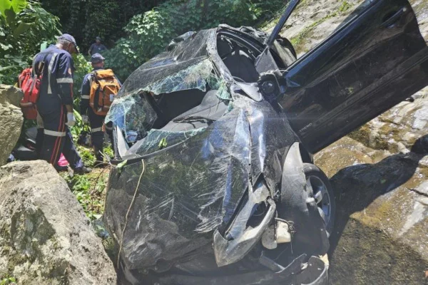 Carro preto com vidro dianteiro destruído está caído entre pedras de ribanceira