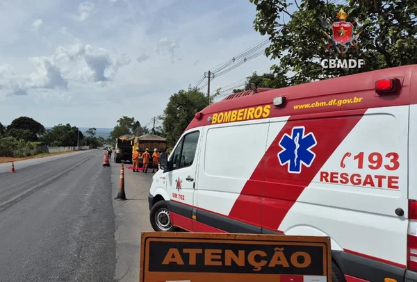 Imagem colorida de uma viatura do CBMDF em frente à cena de um atropelamento; a vítima está no chão, coberta com um lençol vermelho, enquanto quatro bombeiros fardados de laranja estão ao redor