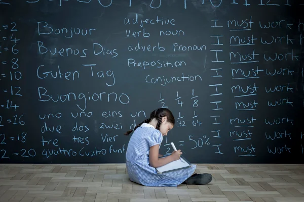 Aluna sentada estudando em frente a um grande quadro-negro com tarefas escolares escritas a giz. Aprendendo outro idioma, Metrópoles