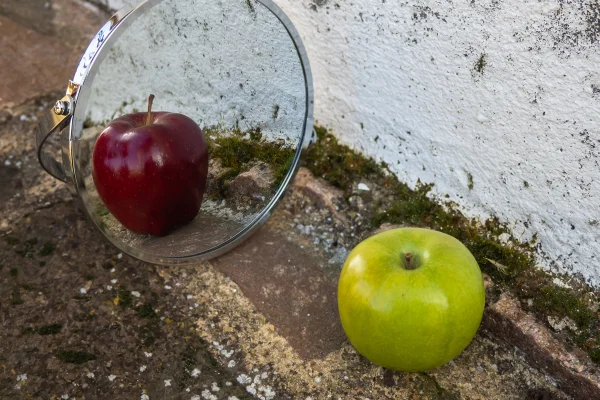 Foto colorida de duas maçãs de cores diferentes e um espelho ao lado de uma parede branca - Met´ropoles.