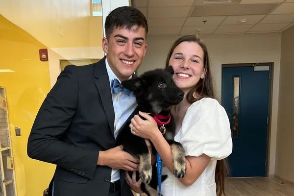 Foto colorida de um casal segurando um cachorro - Metrópoles
