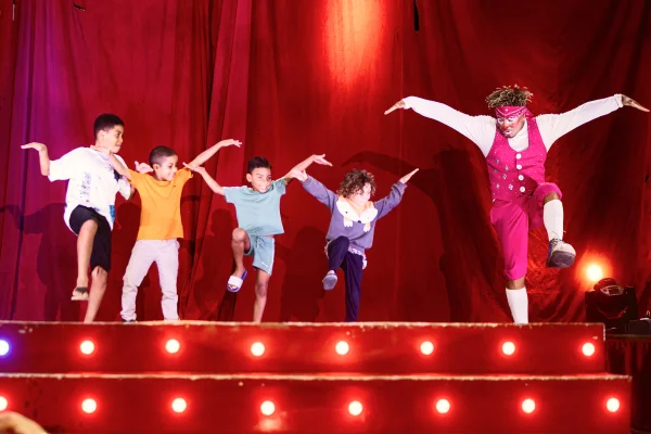 Imagem colorida de um palco onde um palhaço vestido de macacão rosa faz um número de circo com quatro crianças da plateia