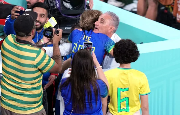 Homem branco, o técnico Tite, usando uma camisa social e no meio da torcida enquanto abraça sua esposa. Uma mulher branca e de cabelo curto vestindo uma blusa azul da Seleção Brasileira escrita "Tite 2022", outras pessoas os cercam enquanto tiram fotos