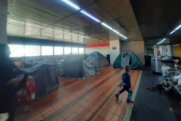 Imagem colorida mostra barracas montadas em corredor do aeroporto de guarulhos. Duas crianças estão na imagem, uma de costas, andando, e a outra olhando para a primeira. Uma mulher de hijab observa a cena na lateral - Metrópoles