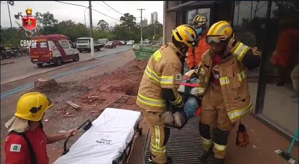 Bombeiros militares levam homem imobilizado em prancha até maca. No fundo, um prédio em obra. Foto colorida